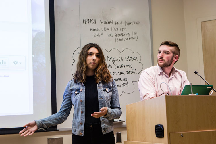 A student presents in front of a screen and whiteboard with a classmate at the podium next to her.