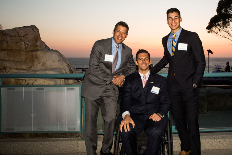 Two gentlemen flank a third guest sitting in a wheelchair with the ocean in the background.