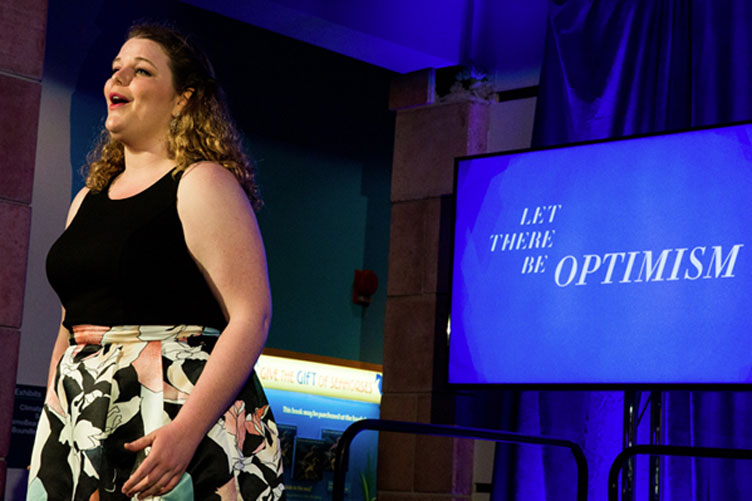 Solo vocalist performs onstage with the segment title “Let There Be Optimism” on the screen behind her.