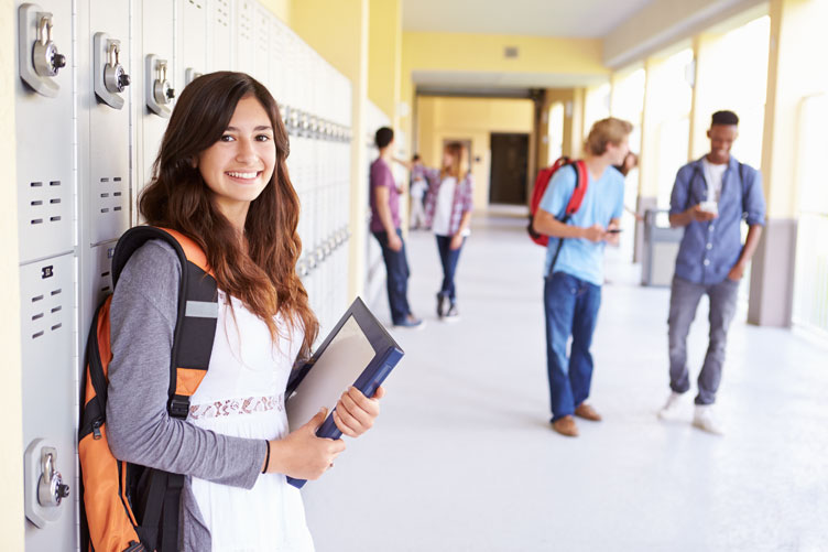 Young female student standing against lockers wearing backpack and with books in hand.