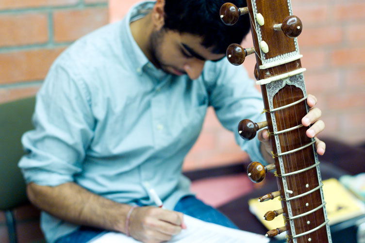 Student holds a stringed instrument in front of him while taking notes in a notebook on his lap.