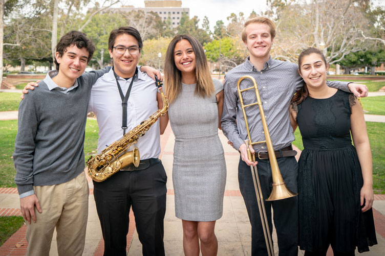 Five student musicians pose for a photo on campus, showing a saxophone and trombone.