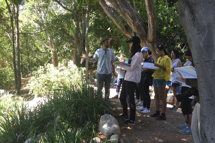 Instructor speaks to students holding notebooks and observing the Mildred E. Mathias Botanical Garden.