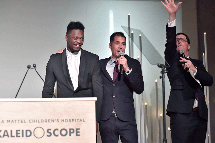 Three men share the stage; one speaks into a microphone and one gestures with his arm raised.