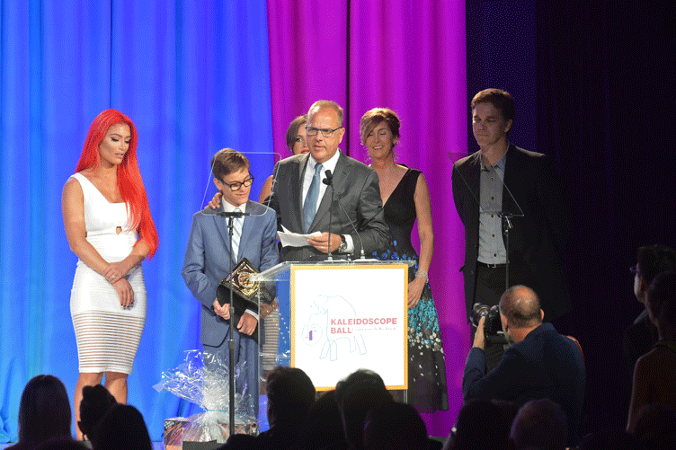 Child recipient and award presenters on stage behind a podium reading “Kaleidoscope Ball.”