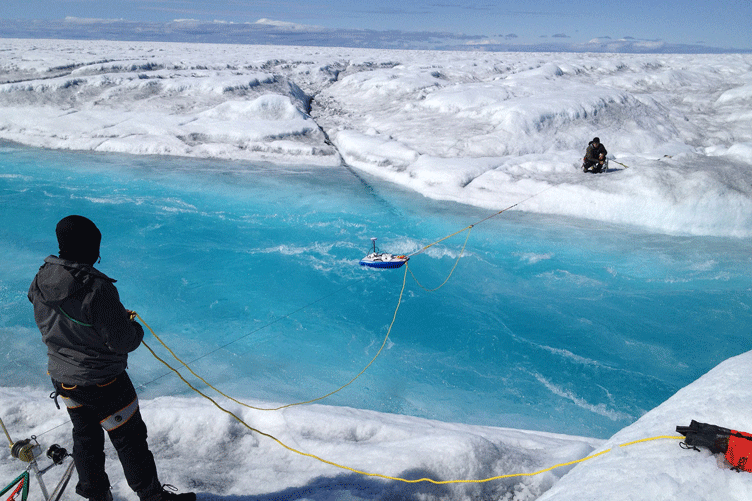 Standing on opposite banks of an icy river, researchers control a floating instrument via rope and wire.