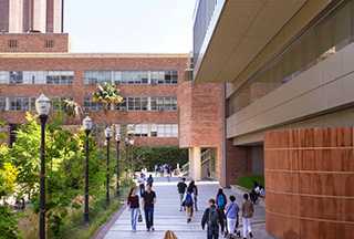 Exterior shot of the brick and stone facade of the NanoSystems Institute building on right, students walking on sidewalk in middle, and a row of trees on the left side