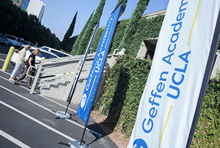 Three free-standing flags showing the Geffen Academy at UCLA logo stand outside the entrance to the ivy covered building