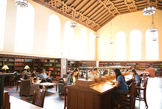 Students studying in a large study space, multiple bookcases filled with books line the walls while a vaulted ceiling and several tall arched windows create a bright and airy space