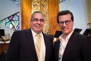 Two white men in glasses, one wearing a dark suit and striped yellow tie and the other wearing a dark blazer with no tie, smile at the camera.