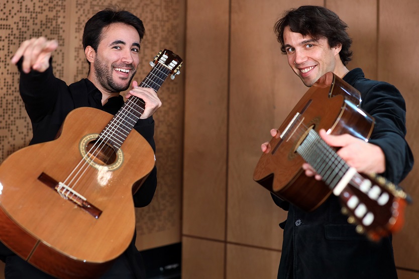 Two dark-haired men wearing dark clothing stand holding up guitars and smiling to camera; man on left points to camera