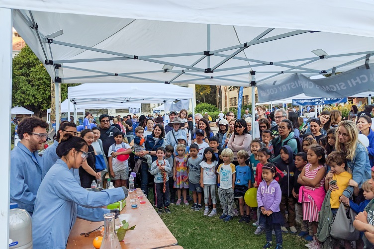 UCLA students on left wear lab coats and goggles and stand behind table to provide a lab demonstration to young children and their families.