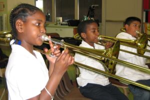 Three students from L.A. schools play the trombone in a classroom; each wears a white shirt and blue jeans.