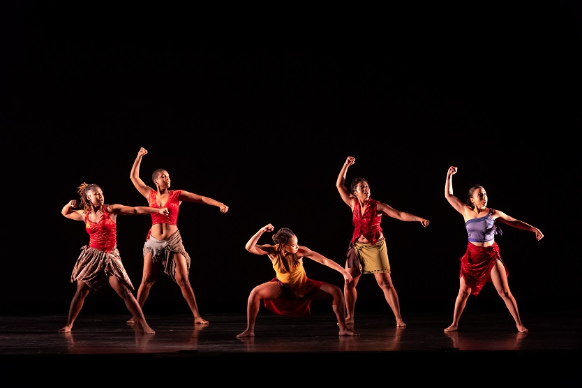 Five dancers in bright tops and wrap skirts hold up their right fists and look to their left; the center dancer is crouched while the others stand.