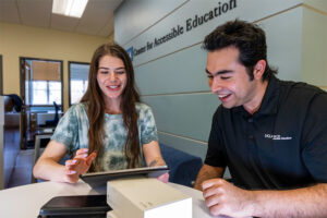 Female student with long medium brown hair looks at a tablet along with a male staff member with short brown hair; both are smiling at what they see, and are sitting under a sign that says "UCLA Center for Accessible Education"