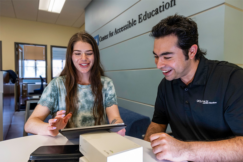 Female student with long medium brown hair looks at a tablet along with a male staff member with short brown hair; both are smiling at what they see, and are sitting under a sign that says "UCLA Center for Accessible Education"