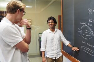 Two male students, one with blonde hair and one with dark, discuss an equation and diagram on a chalkboard.