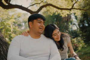 A dark-haired man and woman are seated next to one another in a garden. The woman is leaning into the man and has one arm around his shoulders.