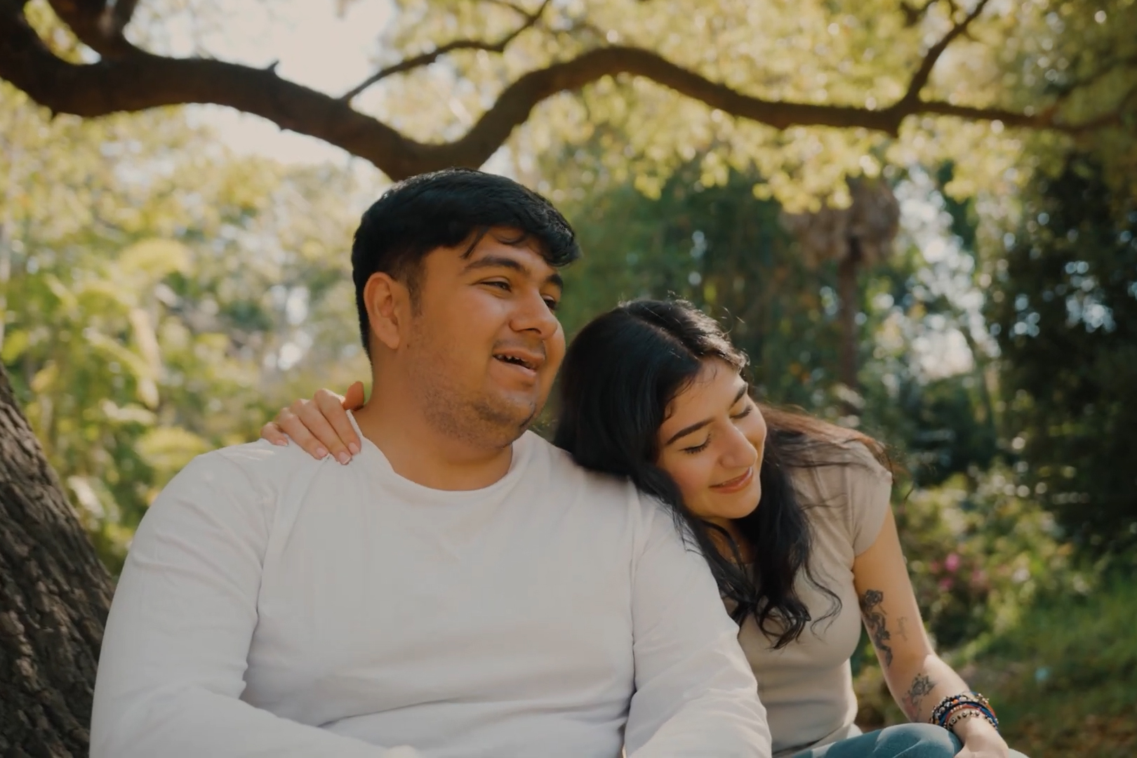 A dark-haired man and woman are seated next to one another in a garden. The woman is leaning into the man and has one arm around his shoulders.