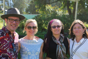 A man in a hat and brightly colored shirt stands to the far left with three women standing to the right of him. All smile at the camera.