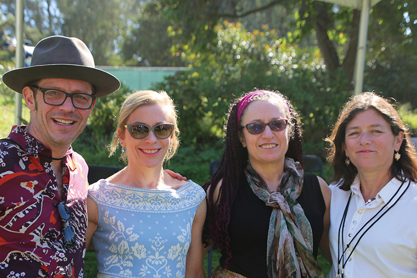 A man in a hat and brightly colored shirt stands to the far left with three women standing to the right of him. All smile at the camera.